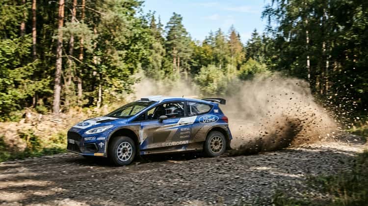 Ein Ford-Rallyefahrzeug fährt auf einer Schotterstrecke durch einen Wald, Steine wirbeln auf, die Szene wirkt dynamisch und kraftvoll.