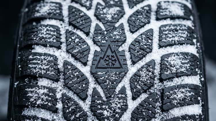 Nahaufnahme eines Winterreifens mit sichtbarem Profil, Schneeflockensymbol und eingefrorenen Schneekristallen in den Lamellen