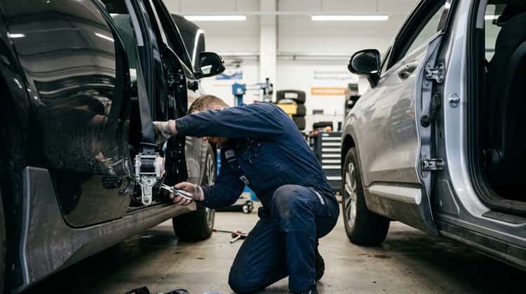 Ein Hyundai Ioniq 6 und ein Hyundai Santa Fe stehen in einer Werkstatt. Ein Mechaniker inspiziert die untere äußere Verankerung der Sicherheitsgurte im Bereich der Fahrzeugtür.